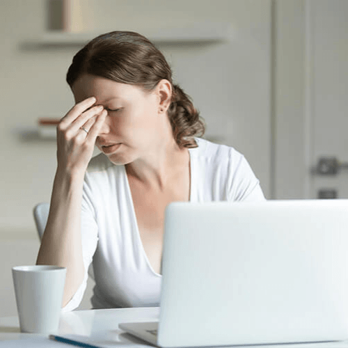 Woman sitting at a laptop holding her forehead while experiencing headache pain and discomfort.